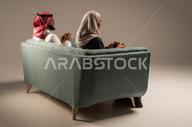  الاستمتاع والتسلية في العطلة، إيماءات وتعابير السرور والتحدي والمتعة، مفهوم الاوقات العائلية السعيده، بورتريه من الخلف لزوجين عربيين سعوديين خليجيين يجلسان على كنبه مريحه يمسكان عصا التحكم يلعبان بألعاب الفيديو، خلفيه رماديه