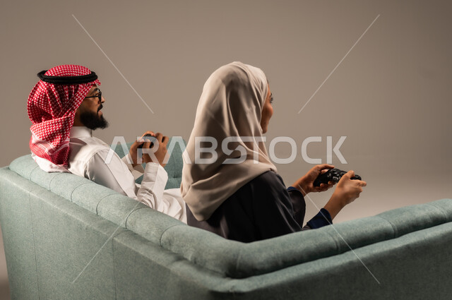  الاستمتاع والتسلية في العطلة، إيماءات وتعابير السرور والتحدي والمتعة، مفهوم الاوقات العائلية السعيده، بورتريه من الخلف لزوجين عربيين سعوديين خليجيين يجلسان على كنبه مريحه يمسكان عصا التحكم يلعبان بألعاب الفيديو، خلفيه رماديه