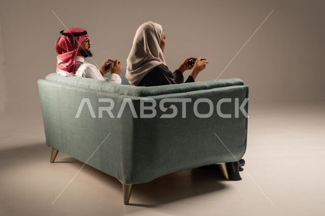  الاستمتاع والتسلية في العطلة، إيماءات وتعابير السرور والتحدي والمتعة، مفهوم الاوقات العائلية السعيده، بورتريه من الخلف لزوجين عربيين سعوديين خليجيين يجلسان على كنبه مريحه يمسكان عصا التحكم يلعبان بألعاب الفيديو، خلفيه رماديه