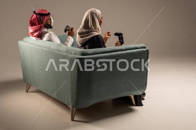  الاستمتاع والتسلية في العطلة، إيماءات وتعابير السرور والتحدي والمتعة، مفهوم الاوقات العائلية السعيده، بورتريه من الخلف لزوجين عربيين سعوديين خليجيين يجلسان على كنبه مريحه يمسكان عصا التحكم يلعبان بألعاب الفيديو، خلفيه رماديه