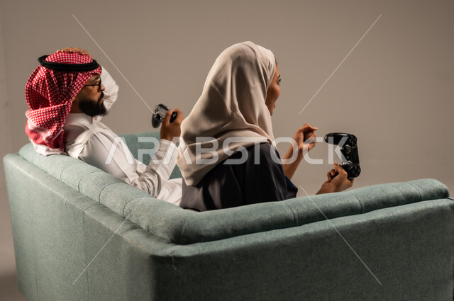  الاستمتاع والتسلية في العطلة، إيماءات وتعابير السرور والتحدي والمتعة، مفهوم الاوقات العائلية السعيده، بورتريه من الخلف لزوجين عربيين سعوديين خليجيين يجلسان على كنبه مريحه يمسكان عصا التحكم يلعبان بألعاب الفيديو، خلفيه رماديه