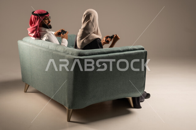 إيماءات وتعابير السرور والتحدي والمتعة، الاستمتاع والتسلية في العطلة، مفهوم الاوقات العائلية السعيده، بورتريه لزوجين عربيين سعوديين خليجيين يجلسان على كنبه مريحه يمسكان عصا التحكم يلعبان بألعاب الفيديو، خلفيه رماديه