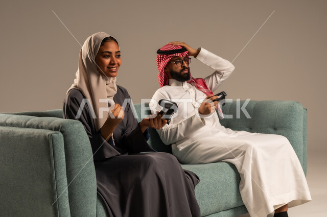 إيماءات وتعابير السرور والتحدي والمتعة، الاستمتاع والتسلية في العطلة، مفهوم الاوقات العائلية السعيده، بورتريه لزوجين عربيين سعوديين خليجيين يجلسان على كنبه مريحه يمسكان عصا التحكم يلعبان بألعاب الفيديو، خلفيه رماديه
