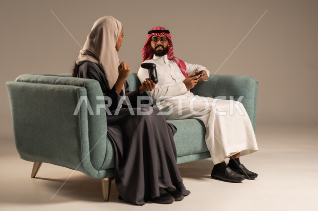 Gestures and expressions of pleasure, challenge and enjoyment, enjoyment and entertainment on vacation, concept of happy family times, portrait of a Saudi Arabian Gulf couple sitting on a comfortable sofa holding a joystick playing video games, gray background
