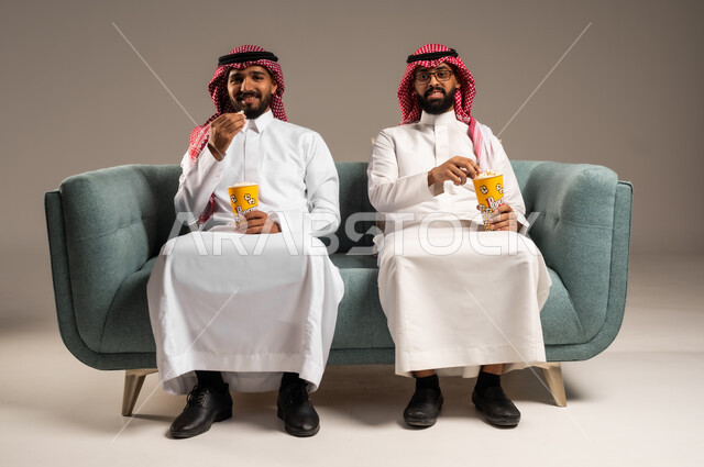مفهوم التسليه والترفيه الجماعي, قضاء وقت ممتع بتناول الفوشار اثناء مشاهده التلفاز، بورتريه لصديقين عربيين خليجيين سعوديين يرتديان الشماغ والثوب التقليدي يجلسان على الأريكه يحملان بيدهما أكواب مليئه بالبوب كورن، تعابير وايماءات السعاده والسرور، خلفية رمادية