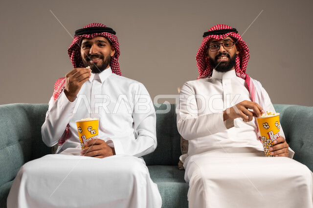 مفهوم التسليه والترفيه الجماعي, قضاء وقت ممتع بتناول الفوشار اثناء مشاهده التلفاز، بورتريه لصديقين عربيين خليجيين سعوديين يرتديان الشماغ والثوب التقليدي يجلسان على الأريكه يحملان بيدهما أكواب مليئه بالبوب كورن، تعابير وايماءات السعاده والسرور، خلفية رمادية