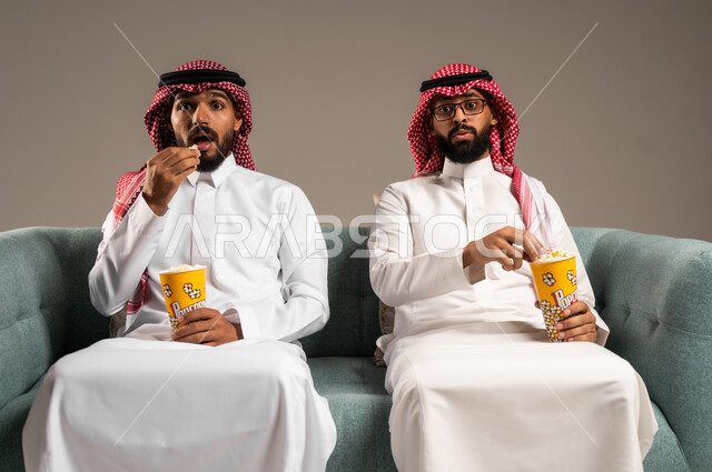 مفهوم التسليه والترفيه الجماعي, قضاء وقت ممتع بتناول الفوشار اثناء مشاهده التلفاز، بورتريه لصديقين عربيين خليجيين سعوديين يرتديان الشماغ والثوب التقليدي يجلسان على الأريكه يحملان بيدهما أكواب مليئه بالبوب كورن، تعابير وايماءات السعاده والسرور، خلفية رمادية