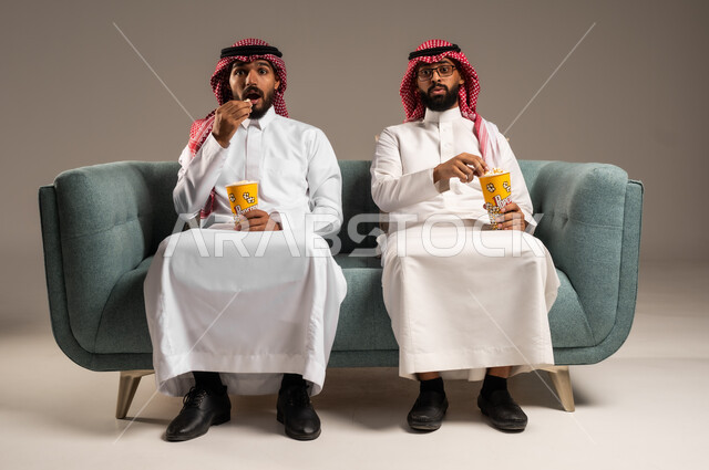 مفهوم التسليه والترفيه الجماعي, قضاء وقت ممتع بتناول الفوشار اثناء مشاهده التلفاز، بورتريه لصديقين عربيين خليجيين سعوديين يرتديان الشماغ والثوب التقليدي يجلسان على الأريكه يحملان بيدهما أكواب مليئه بالبوب كورن، تعابير وايماءات السعاده والسرور، خلفية رمادية