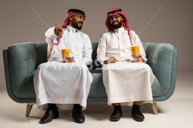 الاشارة الى شيء ما بتعابير السعاده والسرور، مفهوم التسليه والترفيه, قضاء وقت ممتع بتناول الفوشار اثناء مشاهده التلفاز، بورتريه لصديقين عربيين خليجيين سعوديين يرتديان الشماغ والثوب التقليدي يجلسان على الأريكه يحملان بيدهما أكواب مليئه بالبوب كورن، خلفية رمادية