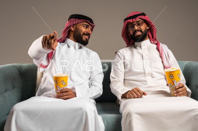 الاشارة الى شيء ما بتعابير السعاده والسرور، مفهوم التسليه والترفيه, قضاء وقت ممتع بتناول الفوشار اثناء مشاهده التلفاز، بورتريه لصديقين عربيين خليجيين سعوديين يرتديان الشماغ والثوب التقليدي يجلسان على الأريكه يحملان بيدهما أكواب مليئه بالبوب كورن، خلفية رمادية