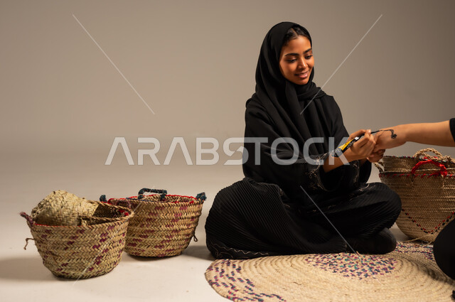 عادات وتقاليد المناسبات والأفراح، بورتريه لإمرأة عربية خليجية سعودية ترتدي الححاب والعباءة تجلس على الأرض بجانب سلل القش تقوم بالرسم والنقش على يد فتاة باستخدام الحناء، مهن تقليدية شعبية نسائية، زخارف يدويه من مواد طبيعيه، الاهتمام بالتجمل والتزيين، خلفية رمادية