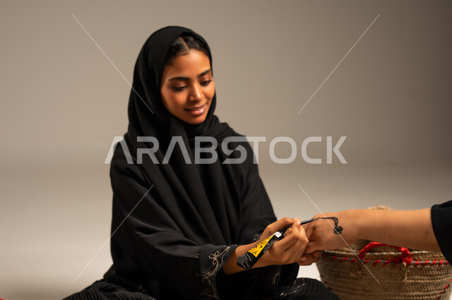Customs and traditions of occasions and weddings, portrait of a Saudi Arabian Gulf woman wearing a hijab and abaya sitting on the ground next to straw baskets drawing and engraving on a girl's hand using henna, traditional popular women's professions, hand decorations from natural materials, interest in beautification and decoration, gray background