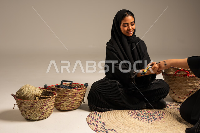 زخارف يدويه من مواد طبيعيه، الاهتمام بالتجمل والتزيين، عادات وتقاليد المناسبات والأفراح، بورتريه لإمرأة عربية خليجية سعودية ترتدي الححاب والعباءة تجلس على الأرض بجانب سلل القش تقوم بالرسم والنقش على يد فتاة باستخدام الحناء، مهن تقليدية شعبية نسائية، خلفية رمادية