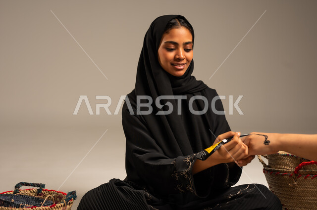 زخارف يدويه من مواد طبيعيه، الاهتمام بالتجمل والتزيين، عادات وتقاليد المناسبات والأفراح، بورتريه لإمرأة عربية خليجية سعودية ترتدي الححاب والعباءة تجلس على الأرض بجانب سلل القش تقوم بالرسم والنقش على يد فتاة باستخدام الحناء، مهن تقليدية شعبية نسائية، خلفية رمادية