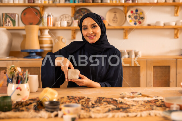 العمل في صناعة الخزفيات، صنع الجرار الفخارية المزخرفة، حرف وصناعات يدوية شعبية من التراث السعودي العريق, اواني خزفية مصنوعة من الطين، صورة مقربة لشابة عربية خليجية سعودية مبتسمة تقوم بصنع الفخار