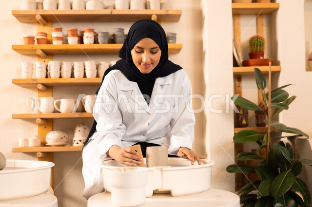العمل في صناعة الخزفيات، صنع الجرار الفخارية المزخرفة، حرف وصناعات يدوية شعبية من التراث السعودي العريق, اواني خزفية مصنوعة من الطين، صورة مقربة لشابة عربية خليجية سعودية مبتسمة تقوم بصنع الفخار