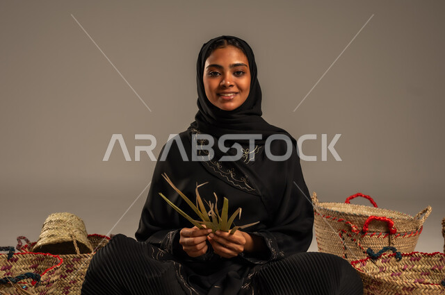 حرف يدوية قديمة تاريخية, مهن نسائية شعبية تقليدية, استخدام أوراق النخيل المجففة في صناعة السلل, منتجات محلية وطنية, التراث والأصالة في السعودية, بورتريه لإمرأة عربية خليجية سعودية ترتدي الحجاب والعباءة تجلس على الارض تقوم بجدل سعائف الخوص، خلفية رمادية