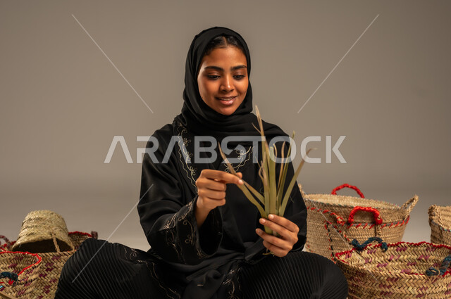 حرف يدوية قديمة تاريخية, مهن نسائية شعبية تقليدية, استخدام أوراق النخيل المجففة في صناعة السلل, منتجات محلية وطنية, التراث والأصالة في السعودية, بورتريه لإمرأة عربية خليجية سعودية ترتدي الحجاب والعباءة تجلس على الارض تقوم بجدل سعائف الخوص، خلفية رمادية
