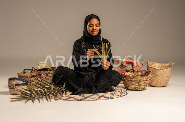 Old historical handicrafts, traditional folk women's professions, using dried palm leaves in basket making, local national products, heritage and authenticity in Saudi Arabia, portrait of a Saudi Arabian Gulf woman wearing a hijab and an abaya sitting on the ground weaving palm fronds, gray background