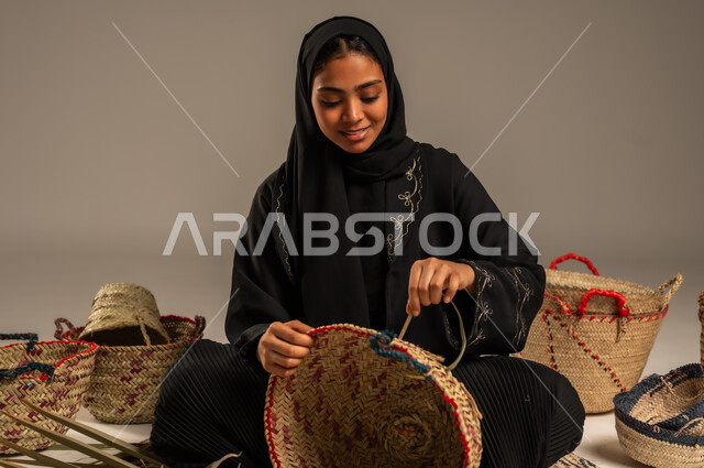 Using dried palm leaves in basket making, local national products, traditional popular women's professions, heritage and authenticity in Saudi Arabia, ancient historical handicrafts, portrait of a Saudi Arabian Gulf woman wearing a hijab and an abaya sitting on the ground weaving palm fronds, gray background