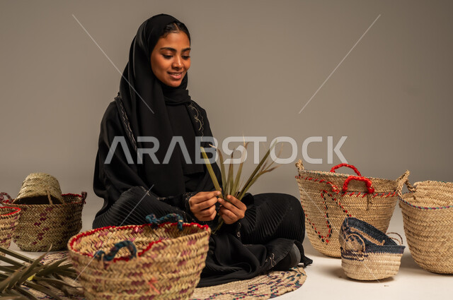 الأصالة والتراث في السعودية, حرف يدوية قديمة تاريخية, مهن نسائية شعبية تقليدية, استخدام أوراق النخيل المجففة في صناعة السلل, منتجات محلية وطنية, بورتريه لإمرأة عربية خليجية سعودية ترتدي الحجاب والعباءة تجلس على الارض تقوم بجدل سعائف الخوص، خلفية رمادية
