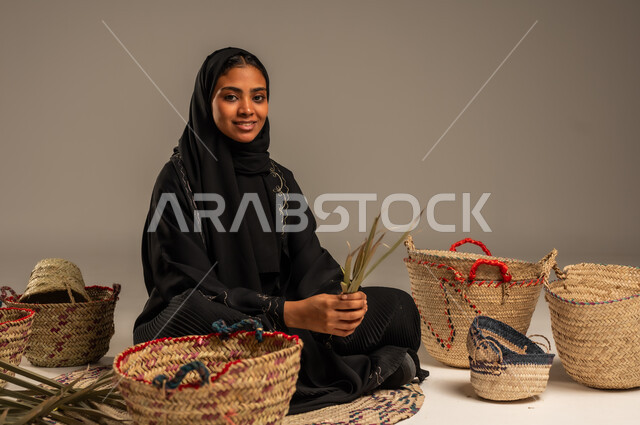 الأصالة والتراث في السعودية, حرف يدوية قديمة تاريخية, مهن نسائية شعبية تقليدية, استخدام أوراق النخيل المجففة في صناعة السلل, منتجات محلية وطنية, بورتريه لإمرأة عربية خليجية سعودية ترتدي الحجاب والعباءة تجلس على الارض تقوم بجدل سعائف الخوص، خلفية رمادية