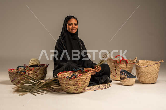 Using dried palm leaves in basket making, local national products, traditional popular women's professions, heritage and authenticity in Saudi Arabia, ancient historical handicrafts, portrait of a Saudi Arabian Gulf woman wearing a hijab and an abaya sitting on the ground weaving palm fronds, gray background
