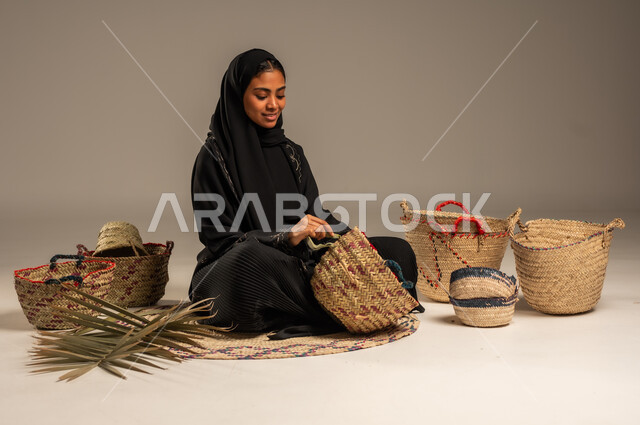 Using dried palm leaves in basket making, local national products, traditional popular women's professions, heritage and authenticity in Saudi Arabia, ancient historical handicrafts, portrait of a Saudi Arabian Gulf woman wearing a hijab and an abaya sitting on the ground weaving palm fronds, gray background