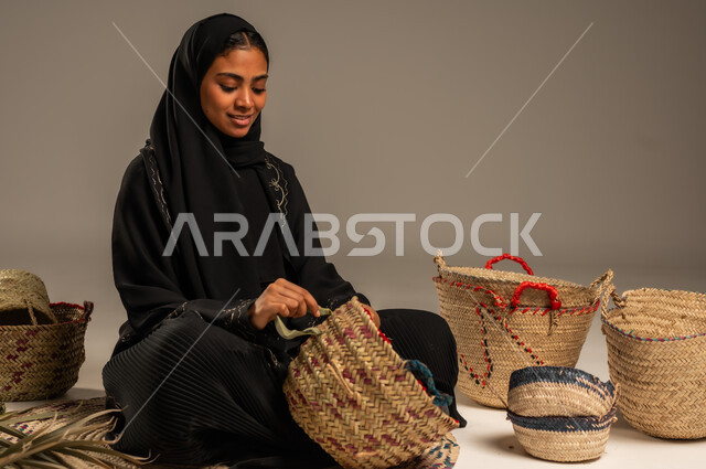 استخدام أوراق النخيل المجففة في صناعة السلل, منتجات محلية وطنية, مهن نسائية شعبية تقليدية, التراث والأصالة في السعودية, حرف يدوية قديمة تاريخية, بورتريه لإمرأة عربية خليجية سعودية ترتدي الحجاب والعباءة تجلس على الارض تقوم بجدل سعائف الخوص، خلفية رمادية