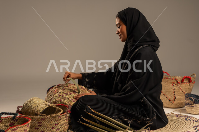 التراث والأصالة في السعودية, حرف يدوية قديمة تاريخية, استخدام أوراق النخيل المجففة في صناعة السلل, منتجات محلية وطنية, مهن نسائية شعبية تقليدية, بورتريه من الجانب لإمرأة عربية خليجية سعودية ترتدي الحجاب والعباءة تجلس على الارض تقوم بجدل سعائف الخوص، خلفية رمادية