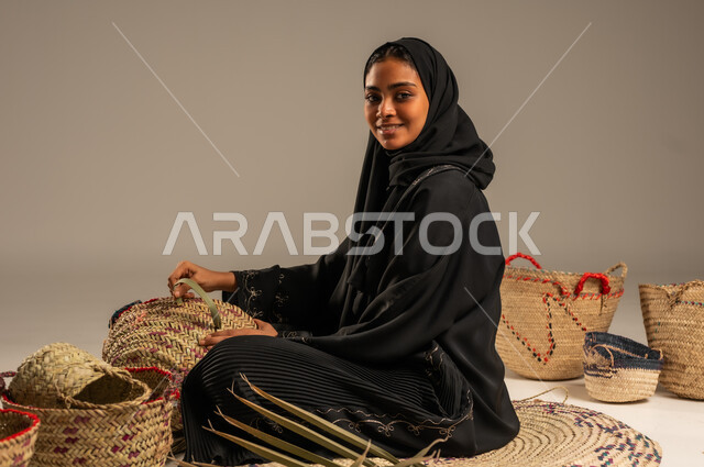 التراث والأصالة في السعودية, حرف يدوية قديمة تاريخية, استخدام أوراق النخيل المجففة في صناعة السلل, منتجات محلية وطنية, مهن نسائية شعبية تقليدية, بورتريه من الجانب لإمرأة عربية خليجية سعودية ترتدي الحجاب والعباءة تجلس على الارض تقوم بجدل سعائف الخوص، خلفية رمادية