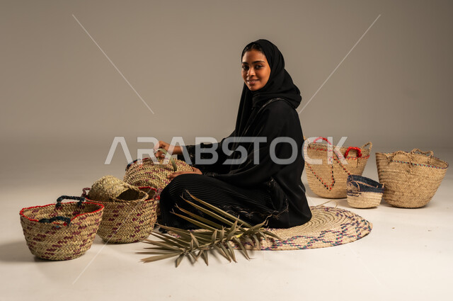 التراث والأصالة في السعودية, حرف يدوية قديمة تاريخية, استخدام أوراق النخيل المجففة في صناعة السلل, منتجات محلية وطنية, مهن نسائية شعبية تقليدية, بورتريه من الجانب لإمرأة عربية خليجية سعودية ترتدي الحجاب والعباءة تجلس على الارض تقوم بجدل سعائف الخوص، خلفية رمادية