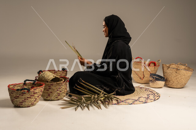 التراث والأصالة في السعودية, حرف يدوية قديمة تاريخية, استخدام أوراق النخيل المجففة في صناعة السلل, منتجات محلية وطنية, مهن نسائية شعبية تقليدية, بورتريه من الجانب لإمرأة عربية خليجية سعودية ترتدي الحجاب والعباءة تجلس على الارض تقوم بجدل سعائف الخوص، خلفية رمادية