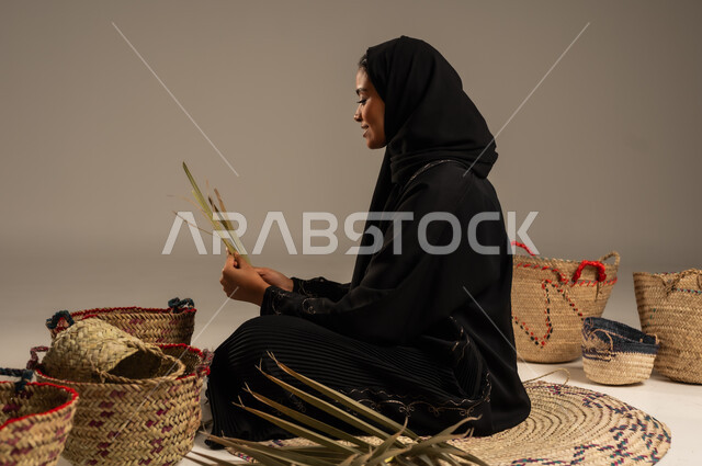 التراث والأصالة في السعودية, حرف يدوية قديمة تاريخية, استخدام أوراق النخيل المجففة في صناعة السلل, منتجات محلية وطنية, مهن نسائية شعبية تقليدية, بورتريه من الجانب لإمرأة عربية خليجية سعودية ترتدي الحجاب والعباءة تجلس على الارض تقوم بجدل سعائف الخوص، خلفية رمادية