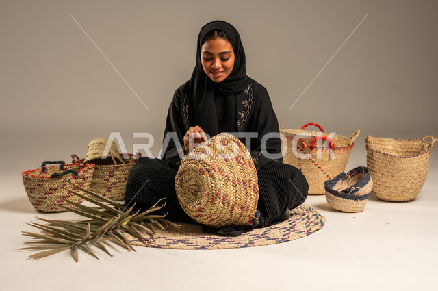 الأصالة والتراث في السعودية, حرف يدوية قديمة تاريخية, مهن نسائية شعبية تقليدية, استخدام أوراق النخيل المجففة في صناعة السلل, منتجات محلية وطنية, بورتريه لإمرأة عربية خليجية سعودية ترتدي الحجاب والعباءة تجلس على الارض تقوم بجدل سعائف الخوص، خلفية رمادية