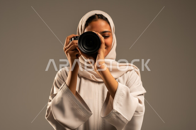 احتراف فن التصوير الفوتوغرافي باستخدام كاميرات رقمية عالية الدقة, وظائف ومهن نسائية سعودية, بورتريه مقرب لإمراة عربية خليجية سعودية ترتدي الحجاب والعباءة تقوم بالتقاط الصورالتذكارية عبر كاميرا فوتوغرافية, خلفية رمادية