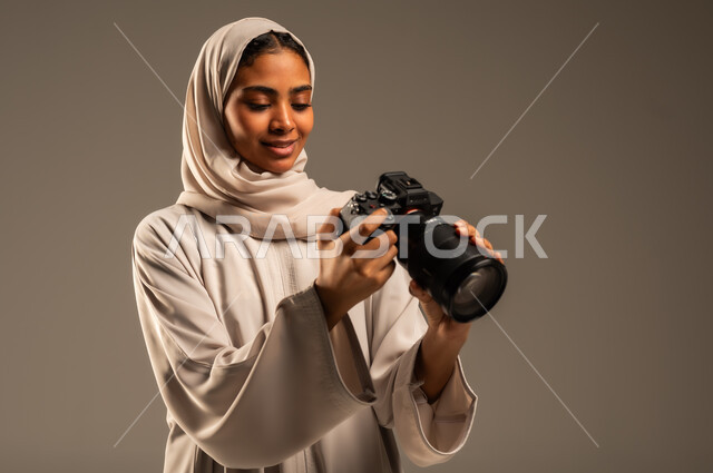 احتراف فن التصوير الفوتوغرافي باستخدام كاميرات رقمية عالية الدقة, وظائف ومهن نسائية سعودية, بورتريه مقرب لإمراة عربية خليجية سعودية ترتدي الحجاب والعباءة تقوم بالتقاط الصورالتذكارية عبر كاميرا فوتوغرافية, خلفية رمادية