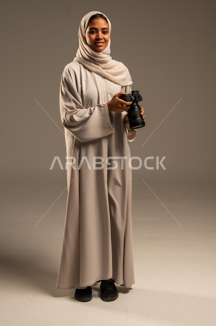 وظائف ومهن نسائية سعودية, احتراف فن التصوير الفوتوغرافي باستخدام كاميرات رقمية عالية الدقة, بورتريه لإمراة عربية خليجية سعودية ترتدي الحجاب والعباءة تقوم بالتقاط الصورالتذكارية عبر كاميرا فوتوغرافية, صورة الجسم بالطول الكامل، خلفية رمادية