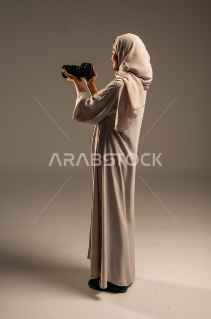 Saudi women's jobs and professions, professional photography using high-resolution digital cameras, portrait of a Saudi Arabian Gulf woman wearing a hijab and abaya taking souvenir photos using a photographic camera, full-length body image, gray background
