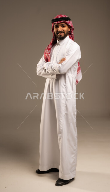 الاعتناء والاهتمام بالمظهر الخارجي، مفهوم الاناقة والرجولة، التقاط صور شخصية للوثائق الرسمية، بورتريه لشاب عربي خليجي سعودي مبتسم يرتدي الثوب التقليدي والشماغ يقف بأيدي متقاطعة ينظر الى الكاميرا بتعابير السرور والثقة بالنفس، الجسم بالطول الكامل، خلفية رمادية