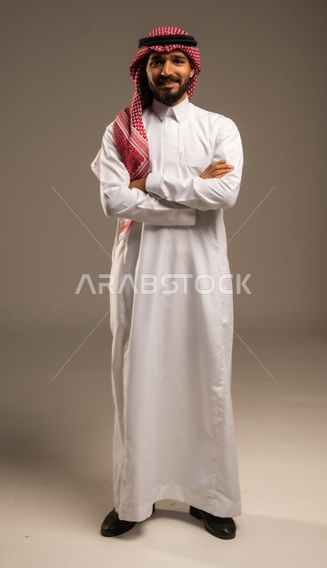 الاعتناء والاهتمام بالمظهر الخارجي، مفهوم الاناقة والرجولة، التقاط صور شخصية للوثائق الرسمية، بورتريه لشاب عربي خليجي سعودي مبتسم يرتدي الثوب التقليدي والشماغ يقف بأيدي متقاطعة ينظر الى الكاميرا بتعابير السرور والثقة بالنفس، الجسم بالطول الكامل، خلفية رمادية