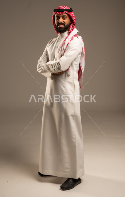 الاعتناء والاهتمام بالمظهر الخارجي، مفهوم الاناقة والرجولة، التقاط صور شخصية للوثائق الرسمية، بورتريه لشاب عربي خليجي سعودي مبتسم يرتدي الثوب التقليدي والشماغ يقف بأيدي متقاطعة ينظر الى الكاميرا بتعابير السرور والثقة بالنفس، الجسم بالطول الكامل، خلفية رمادية