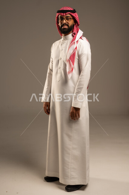 الثقة بالنفس والشعور بالقوة، مفهوم الرجولة والأناقه، الوقوف باستقامة وانضباط، الاهتمام بالمظهر الخارجي، بورتريه لشاب عربي خليجي سعودي مبتسم يرتدي الشماغ والثوب التقليدي يضع يديه على جانبه و ينظر إلى الكاميرا بإيماءات السرور، صورة الجسم بالطول الكامل، خلفية رمادية