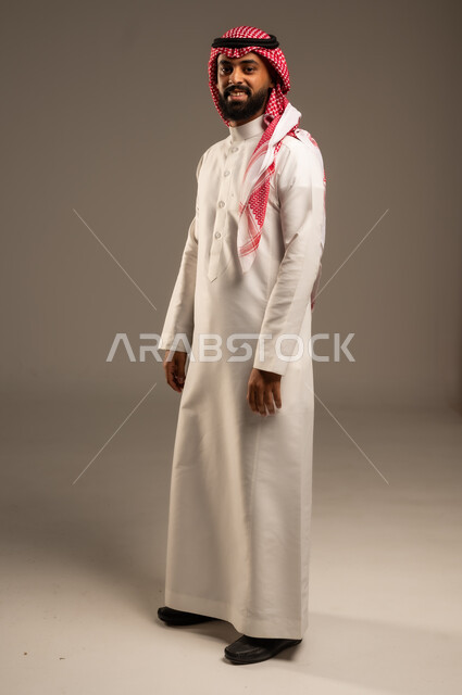 Self-confidence and feeling of strength, concept of masculinity and elegance, standing straight and disciplined, taking care of one's appearance, portrait of a smiling Saudi Arabian Gulf young man wearing a traditional shemagh and thobe, putting his hands on his side and looking at the camera with gestures of pleasure, full-length body image, gray background