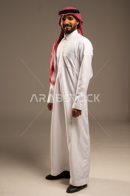 الثقة بالنفس والشعور بالقوة، مفهوم الرجولة والأناقه، الوقوف باستقامة وانضباط، الاهتمام بالمظهر الخارجي، بورتريه لشاب عربي خليجي سعودي مبتسم يرتدي الشماغ والثوب التقليدي يضع يديه على جانبه و ينظر إلى الكاميرا بإيماءات السرور، صورة الجسم بالطول الكامل، خلفية رمادية