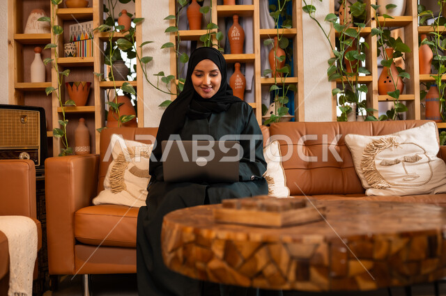 مهن ووظائف نسائية مكتبية، مفهوم إدارة شؤون الأعمال وتنظيمها عبر الاجهزة الحديثة، صورة مقربة لامرأة عربية خليجية سعودية محجبة مبتسمة ترتدي العباءة السوداء تجلس في  محل لبيع الهدايا التذكارية التقليدية تقوم بالعمل على الحاسوب المحمول 