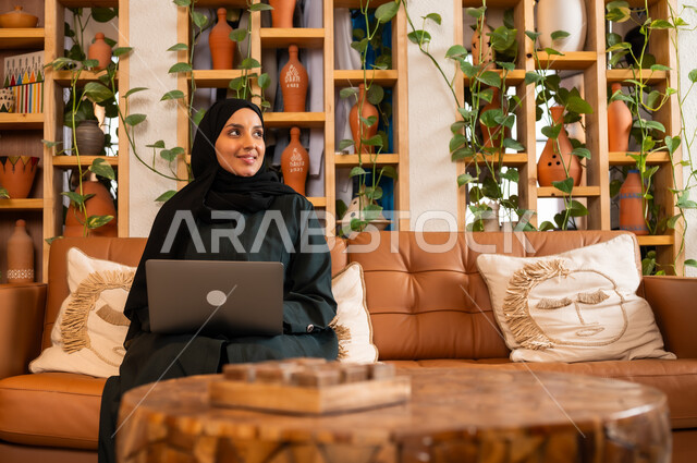 مهن ووظائف نسائية مكتبية، مفهوم إدارة شؤون الأعمال وتنظيمها عبر الاجهزة الحديثة، صورة مقربة لامرأة عربية خليجية سعودية محجبة مبتسمة ترتدي العباءة السوداء تجلس في  محل لبيع الهدايا التذكارية التقليدية تقوم بالعمل على الحاسوب المحمول 