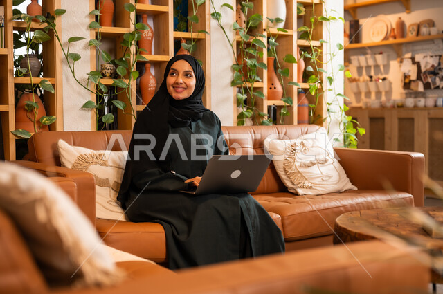 مهن ووظائف نسائية مكتبية، مفهوم إدارة شؤون الأعمال وتنظيمها عبر الاجهزة الحديثة، صورة مقربة لامرأة عربية خليجية سعودية محجبة مبتسمة ترتدي العباءة السوداء تجلس في  محل لبيع الهدايا التذكارية التقليدية تقوم بالعمل على الحاسوب المحمول 