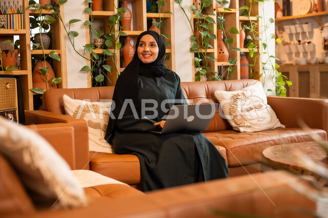 مهن ووظائف نسائية مكتبية، مفهوم إدارة شؤون الأعمال وتنظيمها عبر الاجهزة الحديثة، صورة مقربة لامرأة عربية خليجية سعودية محجبة مبتسمة ترتدي العباءة السوداء تجلس في  محل لبيع الهدايا التذكارية التقليدية تقوم بالعمل على الحاسوب المحمول 