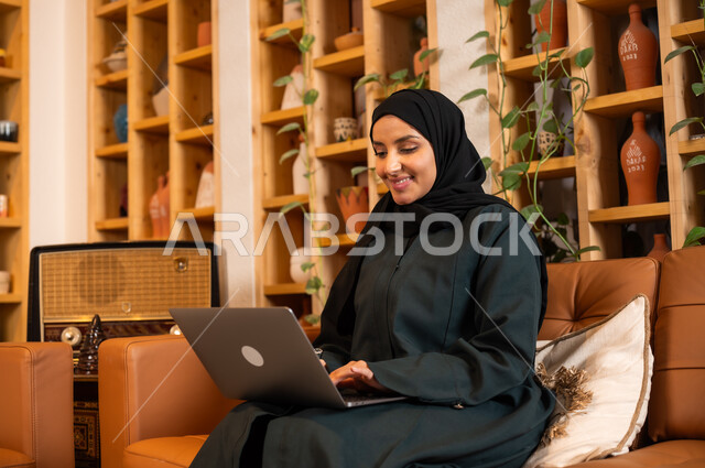 مهن ووظائف نسائية مكتبية، مفهوم إدارة شؤون الأعمال وتنظيمها عبر الاجهزة الحديثة، صورة مقربة لامرأة عربية خليجية سعودية محجبة مبتسمة ترتدي العباءة السوداء تجلس في  محل لبيع الهدايا التذكارية التقليدية تقوم بالعمل على الحاسوب المحمول 
